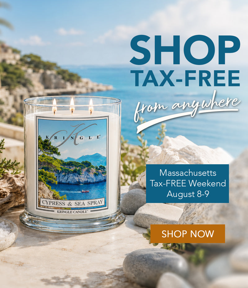 A three-wick scented candle in a clear glass jar sits outdoors on a stone surface with rocks and a coastal landscape in the background. The front label reads KRINGLE and CYPRESS & SEA SPRAY.