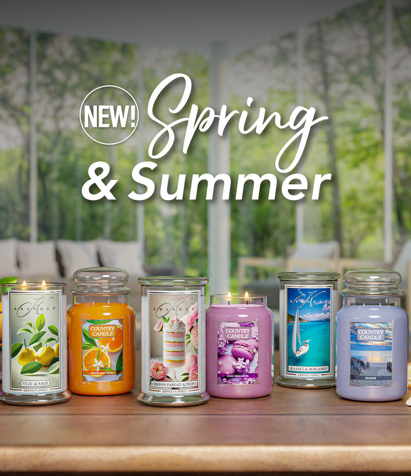 Kringle Candle and Country Candle’s new Spring & Summer collection display. A variety of scented candles in glass jars are showcased on a table with a blurred outdoor background. The candles include scents like Yuzu & Sage, Mandarin Petals, Chiffon Parfait & Peony, Lilac Macaron, Sea Salt & Bergamot, and Seaside. Bright, summery colors and decorative elements such as macarons, citrus fruits, and seashells complement the scene.