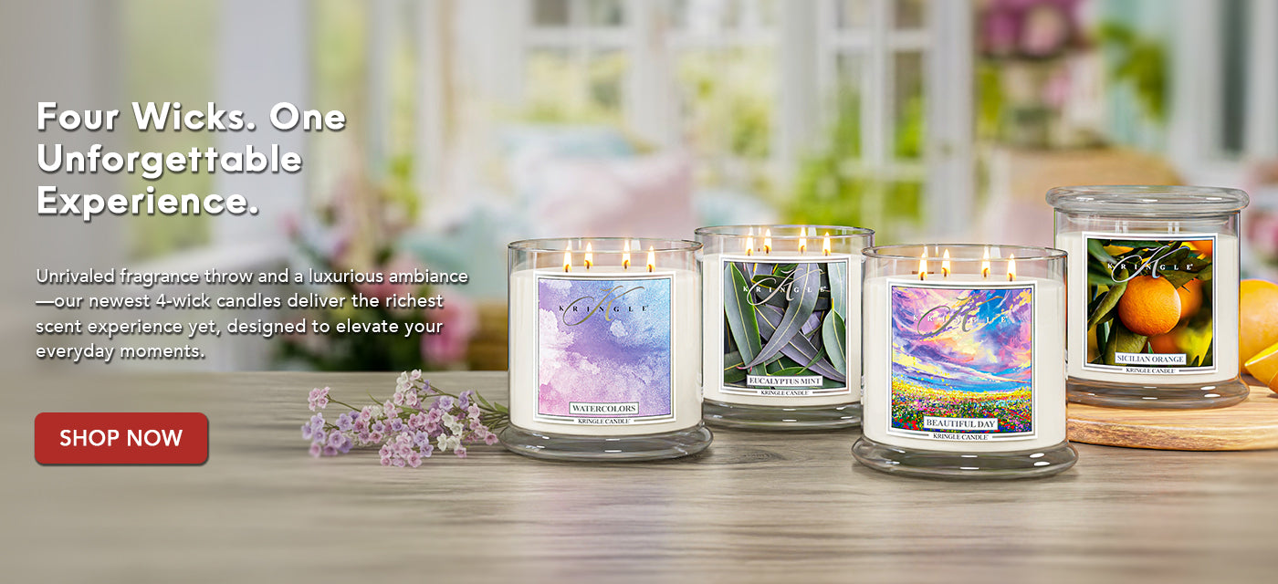 Four wick candles with colorful labels on a wooden surface, with a blurred background of flowers and products.