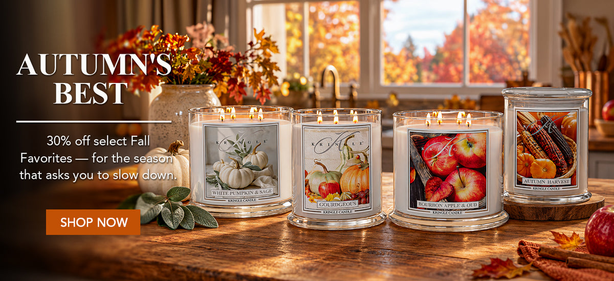 Four lit scented candles in clear glass jars with autumn-themed labels are displayed on a wooden table in a warmly lit kitchen. The labels depict pumpkins, apples, and corn.