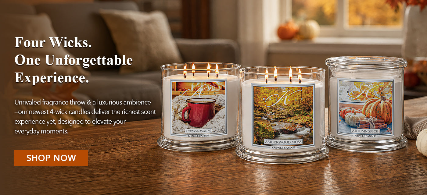 Three lit scented candles in clear glass jars on a wooden table in a cozy living room setting. The left candle shows a red mug in autumn leaves, the middle shows a golden autumn landscape, and the right shows pumpkins and fall foliage.