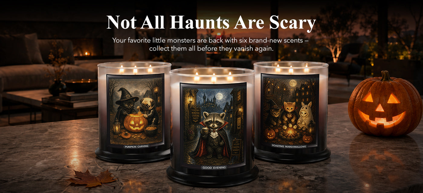 Three lit candles in clear glass jars with black bases are displayed on a dark countertop in a cozy dimly lit room. The labels feature Halloween-themed illustrations including a raccoon, cat, and pumpkin.