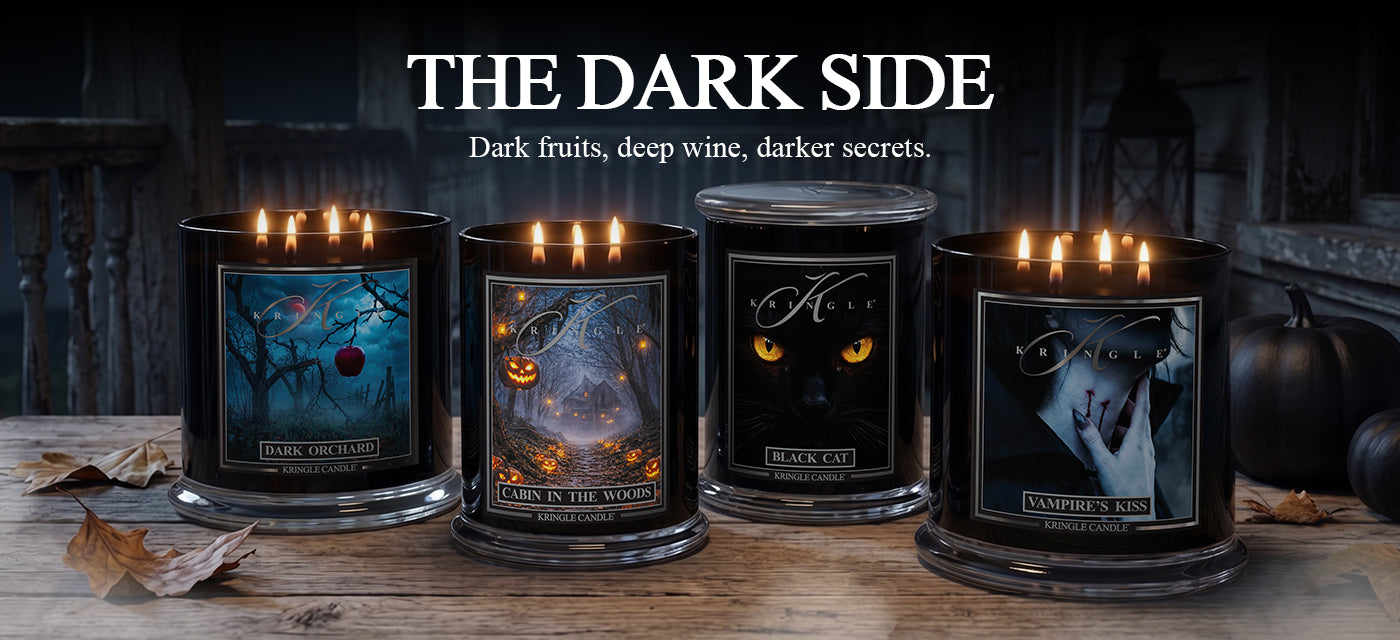 Four lit black glass candles with Halloween-themed labels are displayed on a rustic wooden surface. The candles feature gothic artwork including a blue haunted tree, a pumpkin patch, black cat eyes, and a dark portrait.