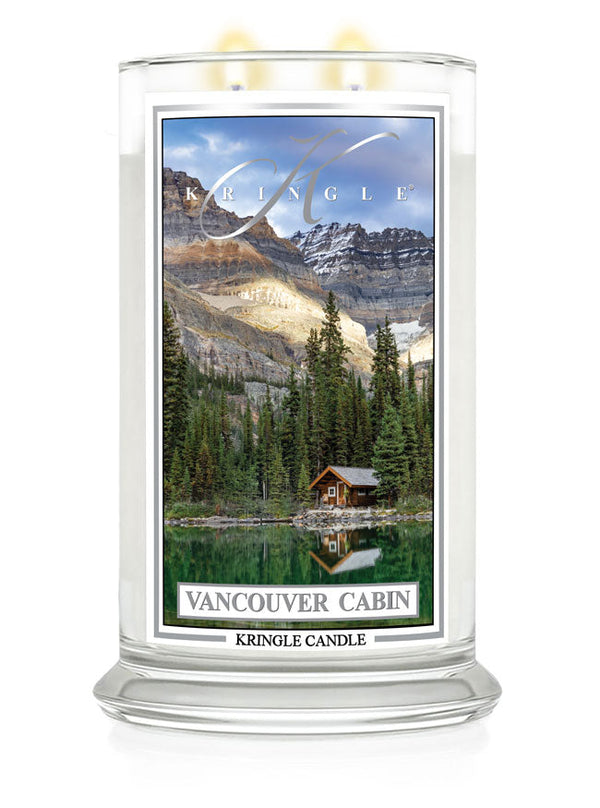 Lit Vancouver Cabin 2-wick candle. Label shows a cabin by a forest and lake with mountains in the background.
