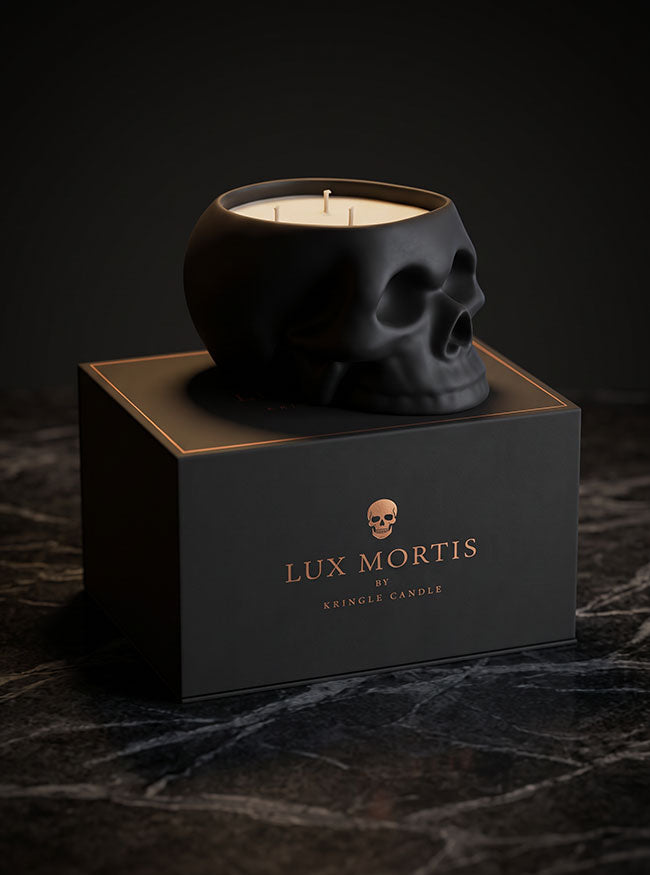 A black skull-shaped cringle candle sits on top of a black branded box labeled Lux Mortis. The candle wax is visible inside the skull-shaped vessel, and the box features gold lettering and a small skull icon.