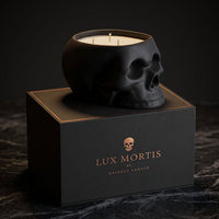 A black skull-shaped cringle candle sits on top of a black branded box labeled Lux Mortis. The candle wax is visible inside the skull-shaped vessel, and the box features gold lettering and a small skull icon.