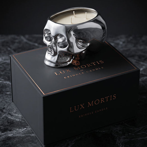 A silver skull-shaped crinkle candle sits on top of a black Lux Mortis box on a dark marble surface against a black background.
