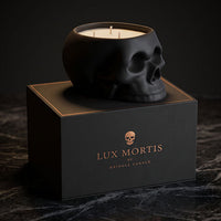 A black skull-shaped cringle candle sits on top of a black branded box labeled Lux Mortis. The candle wax is visible inside the skull-shaped vessel, and the box features gold lettering and a small skull icon.