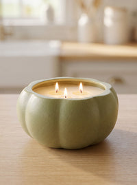 A light sage green pumpkin-shaped candle holder containing two lit candles, set on a light wood table with a softly blurred kitchen background.