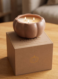 A lit two-wick candle in a ribbed pumpkin-shaped ceramic holder sits on top of its branded kraft gift box on a wooden table. The box reads 'Pumpkin Collection' and features a gold pumpkin outline.