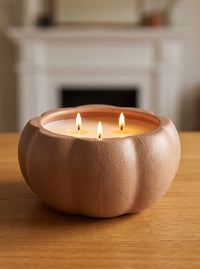 A three-wick candle in a matte terracotta pumpkin-shaped holder, lit with three flames, sitting on a wooden table with a blurred fireplace in the background.