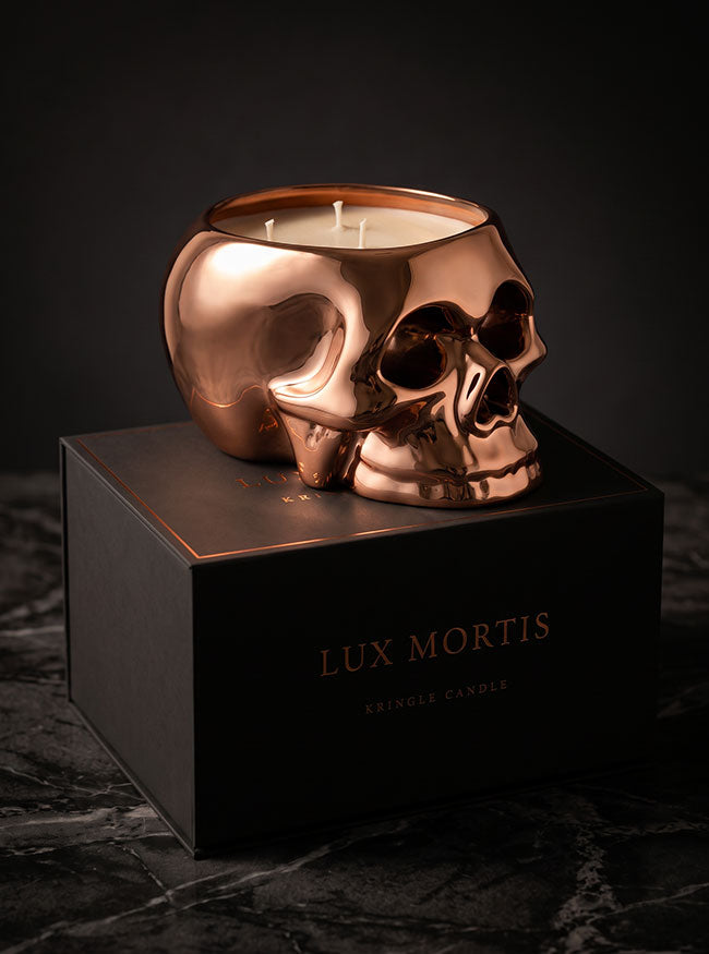 A metallic copper skull-shaped candle holder filled with white wax, displayed on top of a black box labeled 'LUX MORTIS' against a dark background.