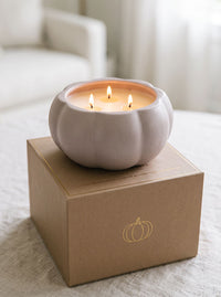 A lit three-wick candle in a ribbed, pumpkin-shaped ceramic holder sits on top of a kraft paper gift box with a gold pumpkin outline, set on a soft beige fabric surface.