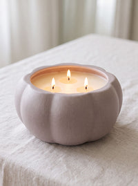 A three-wick candle in a matte, pumpkin-shaped ceramic holder with a warm cream wax, set on a light beige linen tablecloth with a softly blurred background.