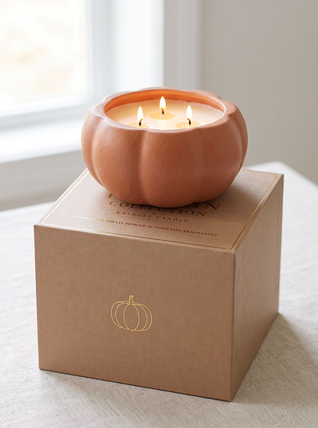 A lit terracotta pumpkin-shaped candle sits on top of a kraft paper gift box with a gold pumpkin outline and the words 'Pumpkin Collection Scented Candle.' The scene is photographed indoors near a window on a light fabric surface.