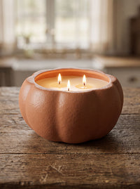 A terracotta pumpkin-shaped candle holder with two lit candles inside, set on a rustic wooden table with a softly blurred indoor background.