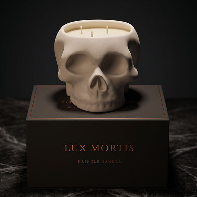 A skull-shaped candle in an off-white ceramic finish sits on top of a dark brown box labeled 'LUX MORTIS' and 'KRINGLE CANDLE'. The scene is photographed on a dark marble surface against a dark background.