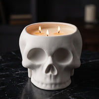A white ceramic skull-shaped candle holder containing three lit beeswax candles, set on a dark marble surface with a dark, moody background.