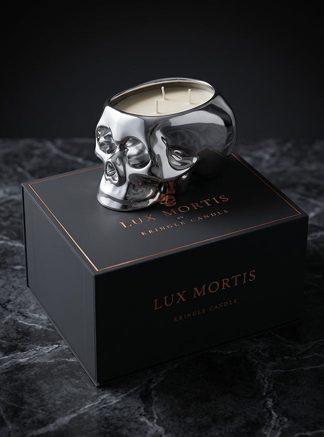 A silver skull-shaped crinkle candle sits on top of a black Lux Mortis box on a dark marble surface against a black background.