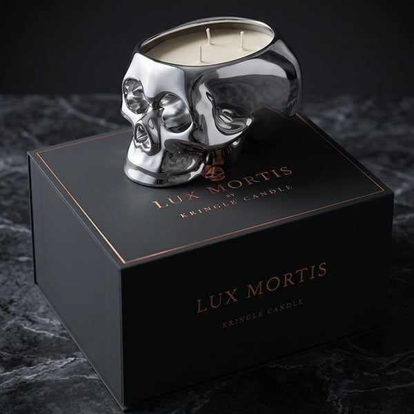 A silver skull-shaped crinkle candle sits on top of a black Lux Mortis box on a dark marble surface against a black background.