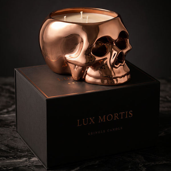 A metallic copper skull-shaped candle holder filled with white wax, displayed on top of a black box labeled 'LUX MORTIS' against a dark background.