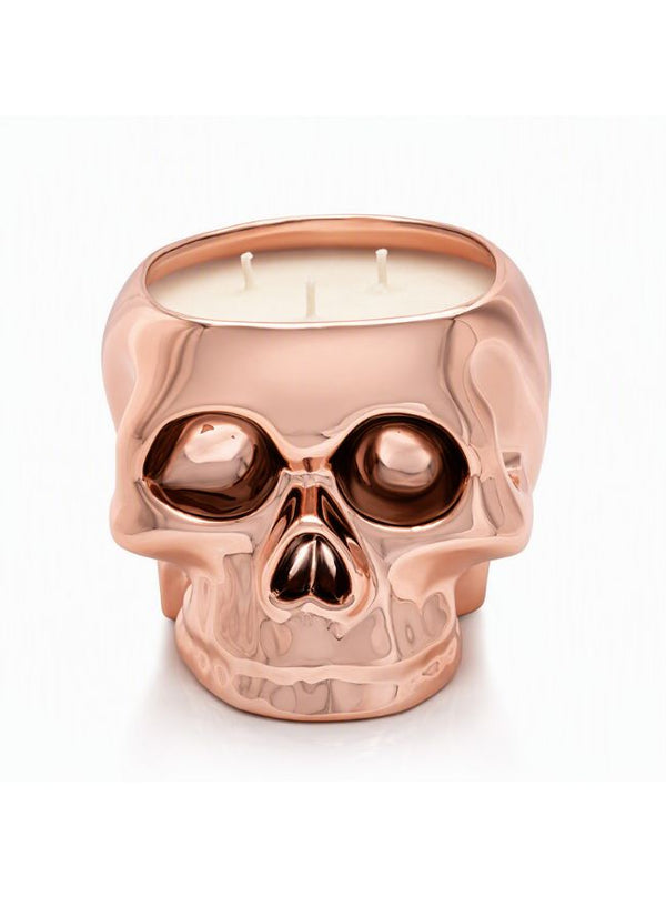 A skull-shaped candle with a reflective rose gold metallic finish, featuring three wicks on a white wax fill, set against a plain white background.