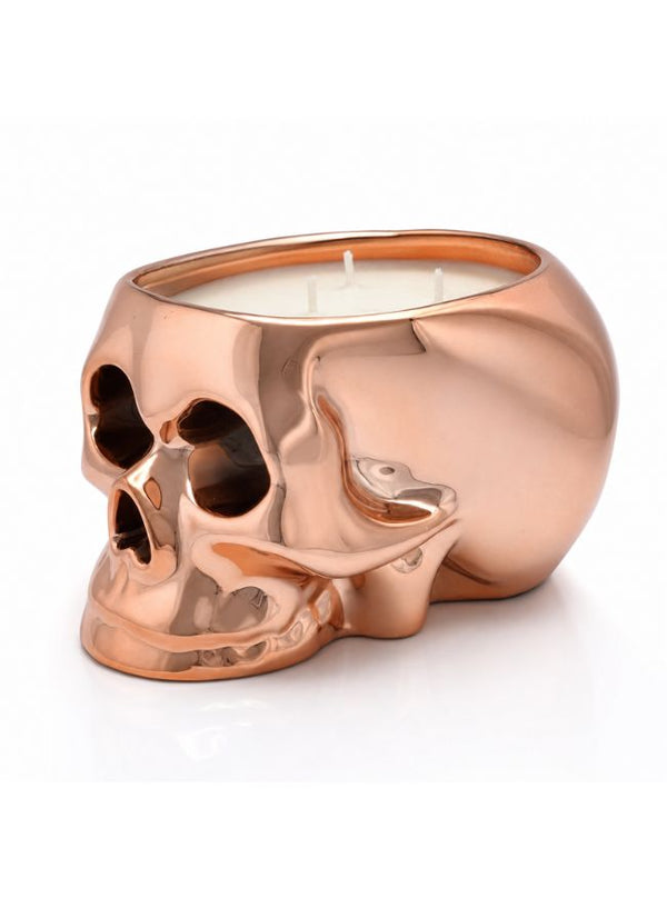 A skull-shaped candle holder in a shiny rose gold metallic finish, filled with white wax and featuring three wicks, set against a plain white background.