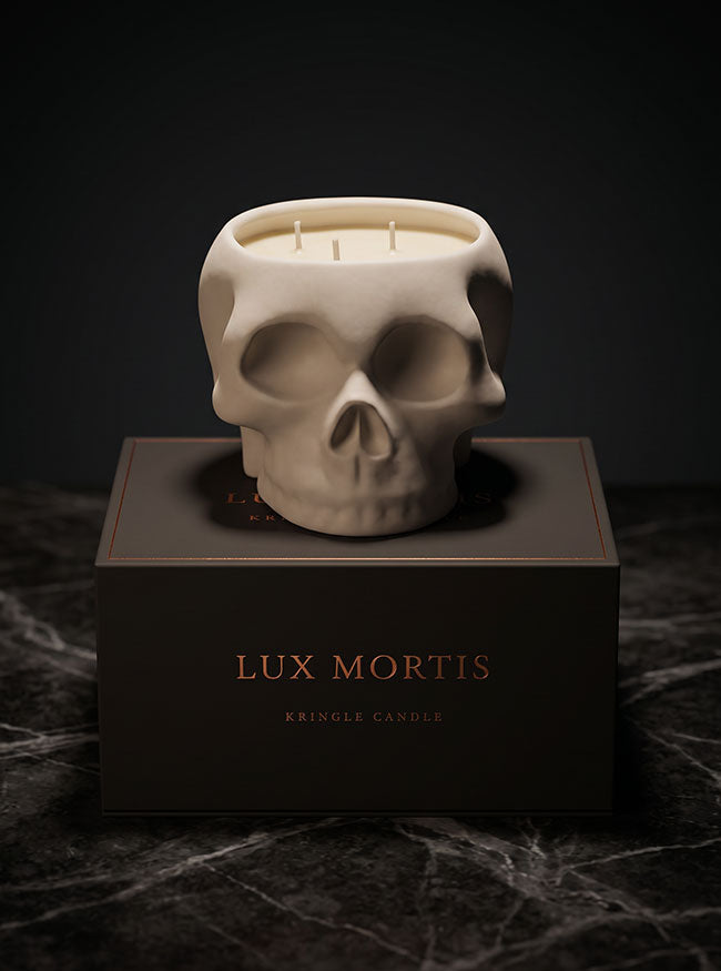 A skull-shaped candle in an off-white ceramic finish sits on top of a dark brown box labeled 'LUX MORTIS' and 'KRINGLE CANDLE'. The scene is photographed on a dark marble surface against a dark background.