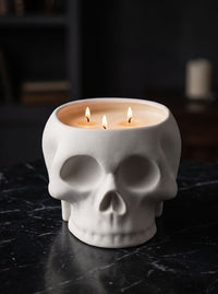 A white ceramic skull-shaped candle holder containing three lit beeswax candles, set on a dark marble surface with a dark, moody background.