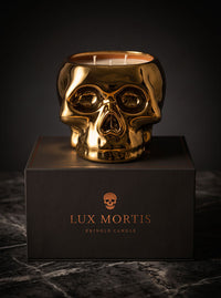 A metallic gold skull-shaped cringle candle sits on top of a black branded box. The candle has two visible wicks and a reflective polished finish, photographed against a dark studio background with a marble surface below.
