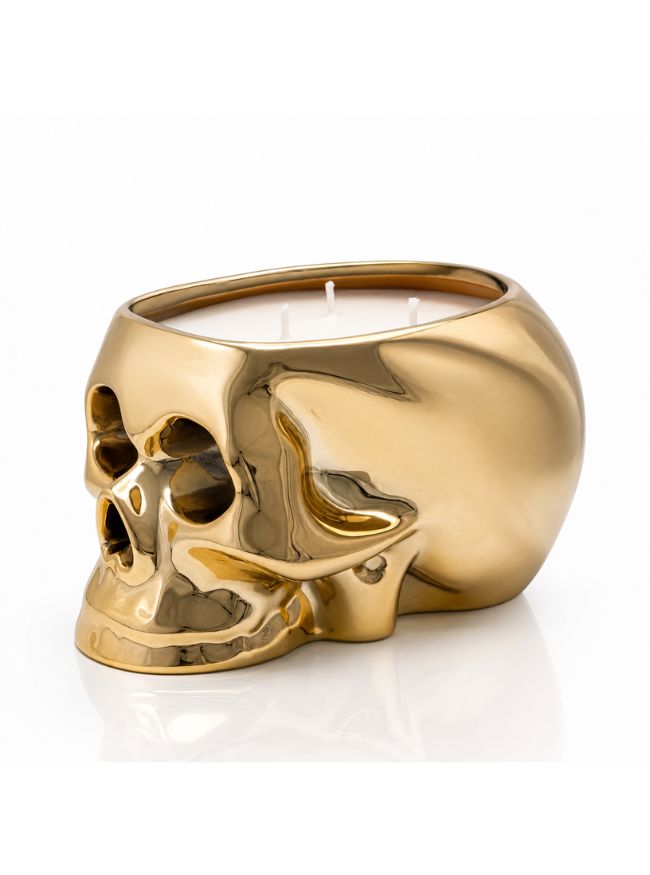 A metallic gold skull-shaped candle holder containing white wax, photographed on a clean white background with a soft reflection below.