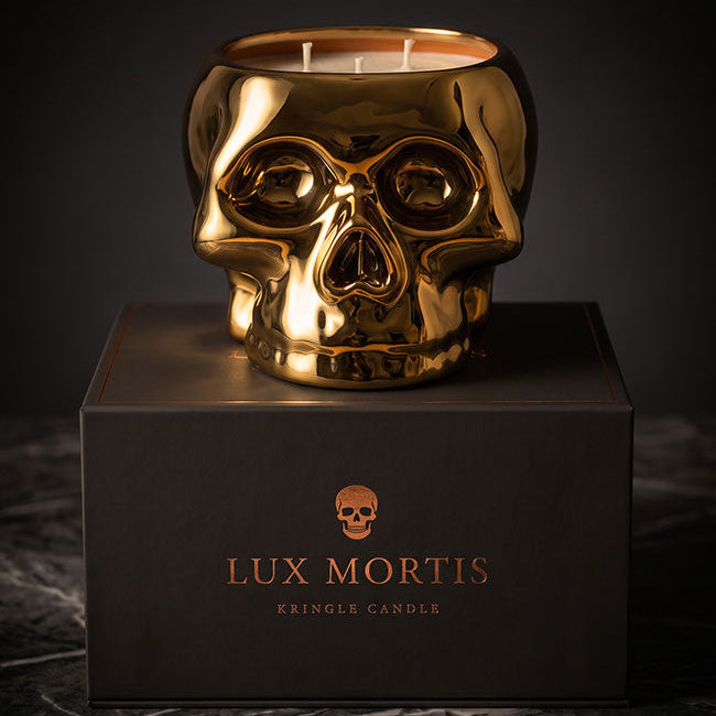 A metallic gold skull-shaped cringle candle sits on top of a black branded box. The candle has two visible wicks and a reflective polished finish, photographed against a dark studio background with a marble surface below.