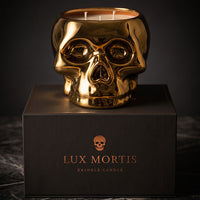 A metallic gold skull-shaped cringle candle sits on top of a black branded box. The candle has two visible wicks and a reflective polished finish, photographed against a dark studio background with a marble surface below.