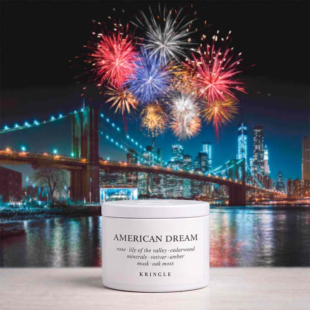 A white jar of KRINGLE American Dream hair product sits on a light surface in front of a nighttime city skyline with fireworks over the water and bridge.