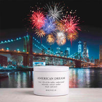 A white jar of KRINGLE American Dream hair product sits on a light surface in front of a nighttime city skyline with fireworks over the water and bridge.