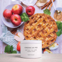 A white jar candle labeled 'AMERICAN PIE' by Kringle, styled on a tabletop with an apple pie, red apples, cinnamon sticks, and green leaves in the background.
