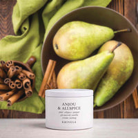 A white scented candle jar labeled 'ANJOU & ALLSPICE' by KRINGLE sits on a light surface in front of a bowl of green pears and cinnamon sticks, with a green cloth draped in the background.