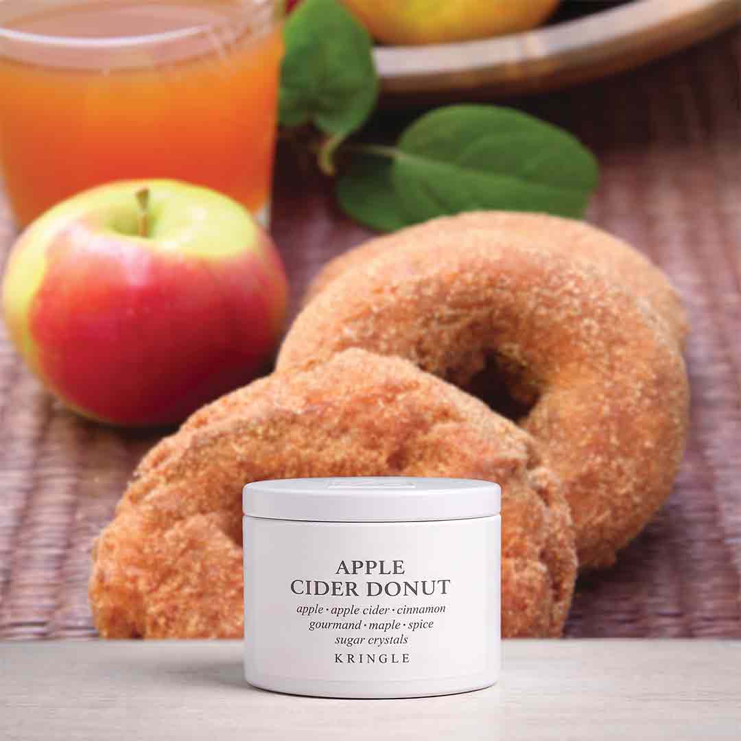 A white jar of Kringle Apple Cider Donut cream sits in front of two sugar-coated donuts on a woven mat, with an apple, a glass of orange juice, and leaves blurred in the background.