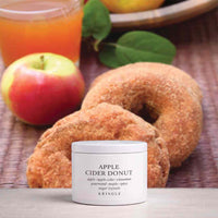 A white jar of Kringle Apple Cider Donut cream sits in front of two sugar-coated donuts on a woven mat, with an apple, a glass of orange juice, and leaves blurred in the background.