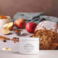 A white cylindrical tin of KRINGLE Apple Rum Cake perfume placed on a light surface, with sliced bread, apples, cinnamon sticks, and a gray cloth in the background.