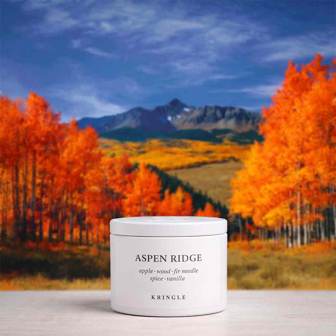 A white cylindrical candle tin labeled 'ASPEN RIDGE' and 'KRINGLE' sits on a light surface in front of a scenic autumn landscape with orange trees, blue sky, and mountains.