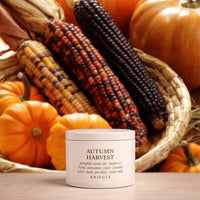 A white jar of KRINGLE 'Autumn Harvest' body butter sits in front of a rustic autumn-themed background featuring a woven basket filled with corn, pumpkins, and dried leaves.