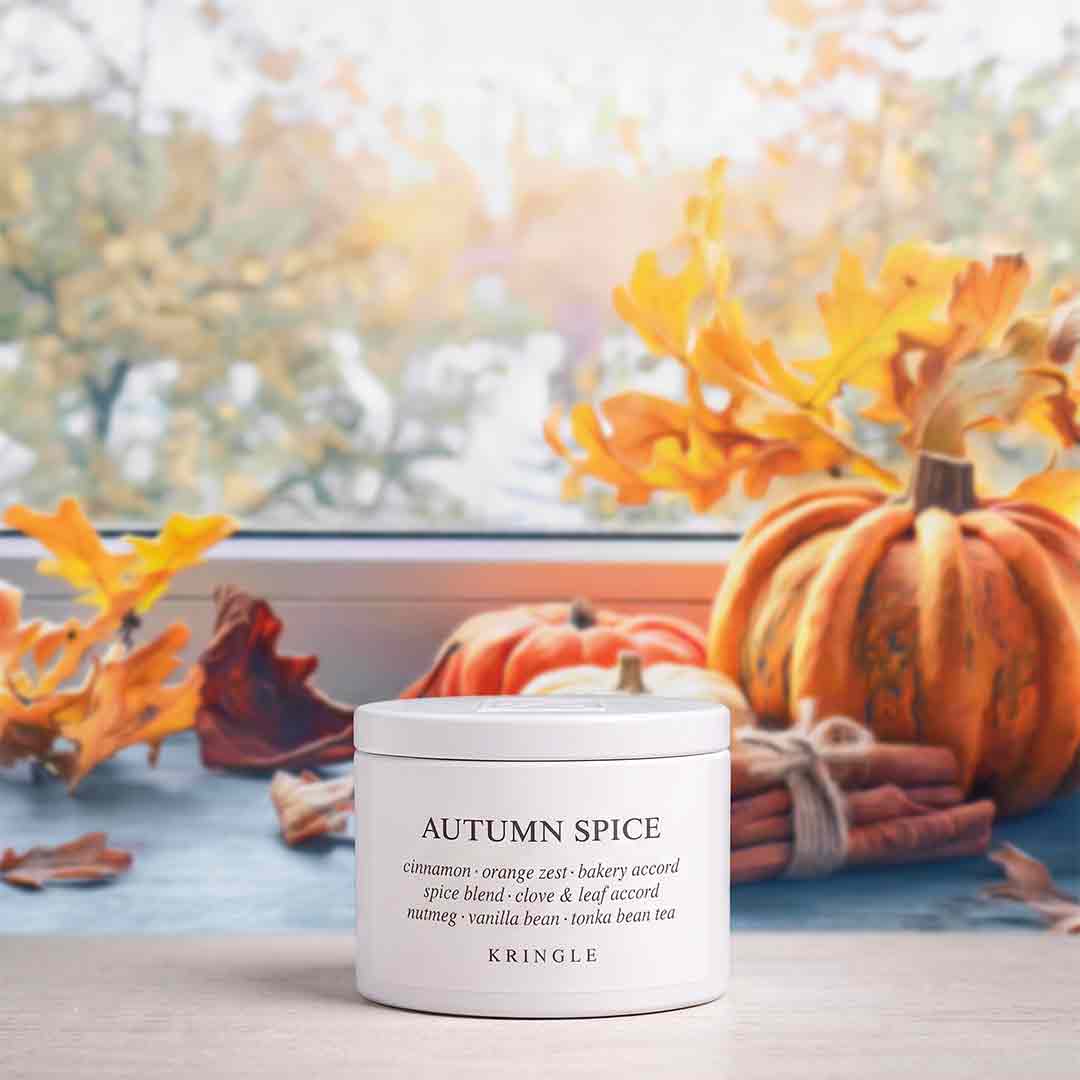 A white jar of KRINGLE Autumn Spice candle sits on a windowsill surrounded by autumn decorations, including orange pumpkins, dried leaves, cinnamon sticks, and a blurred window view of fall trees in the background.
