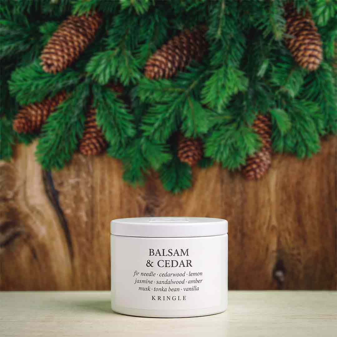 A white jar of Kringle Balsam & Cedar facial mask placed on a light surface, with green pine branches and brown pinecones in the background against a rustic wood backdrop.