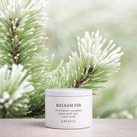 A white jar of KRINGLE Balsam Fir natural body butter sits on a light wooden surface, framed by frosted green pine branches against a soft, blurred light green background.