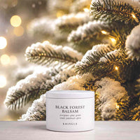 A white jar of KRINGLE Black Forest Balsam placed on a light surface in front of frosted evergreen branches with warm string lights, creating a holiday-themed background.