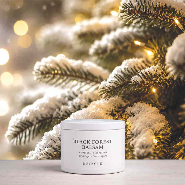 A white jar of KRINGLE Black Forest Balsam placed on a light surface in front of frosted evergreen branches with warm string lights, creating a holiday-themed background.