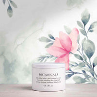A white jar of KRINGLE Botanicals body wash with a white lid, set on a light wooden surface against a watercolor background of green leaves and a pink flower.