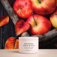 A white cylindrical candle jar labeled 'BOURBON APPLE & OUD' by KRINGLE sits on a light wood surface, with red apples and autumn leaves blurred in the background.