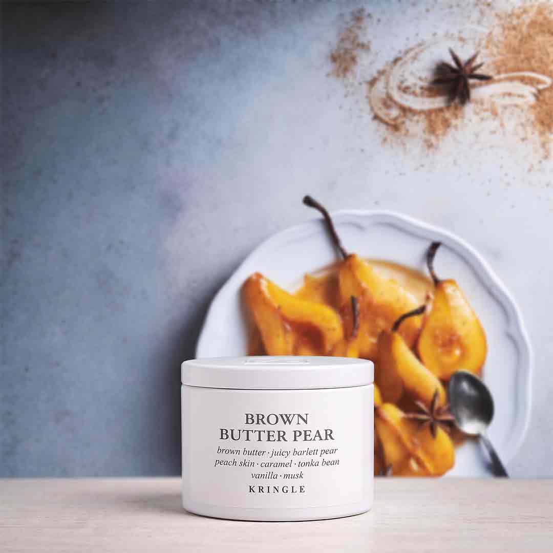 A white jar of KRINGLE Brown Butter Pear facial mask sits on a light wooden surface in front of a white plate of cooked pears with star anise and a spoon, against a textured blue-gray background.